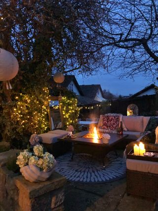 a fire pit in outdoor seating area by night, with lots of garden furniture, potted flowers, candles, and fairy lights