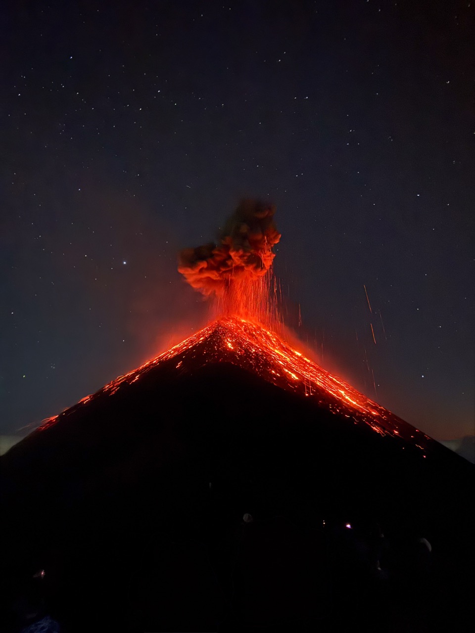 A photograph of a volcano mid eruption at night, with the bright red molten rock contrasting greatly against the dark-shrouded mountainside and the dimly starlit night sky.