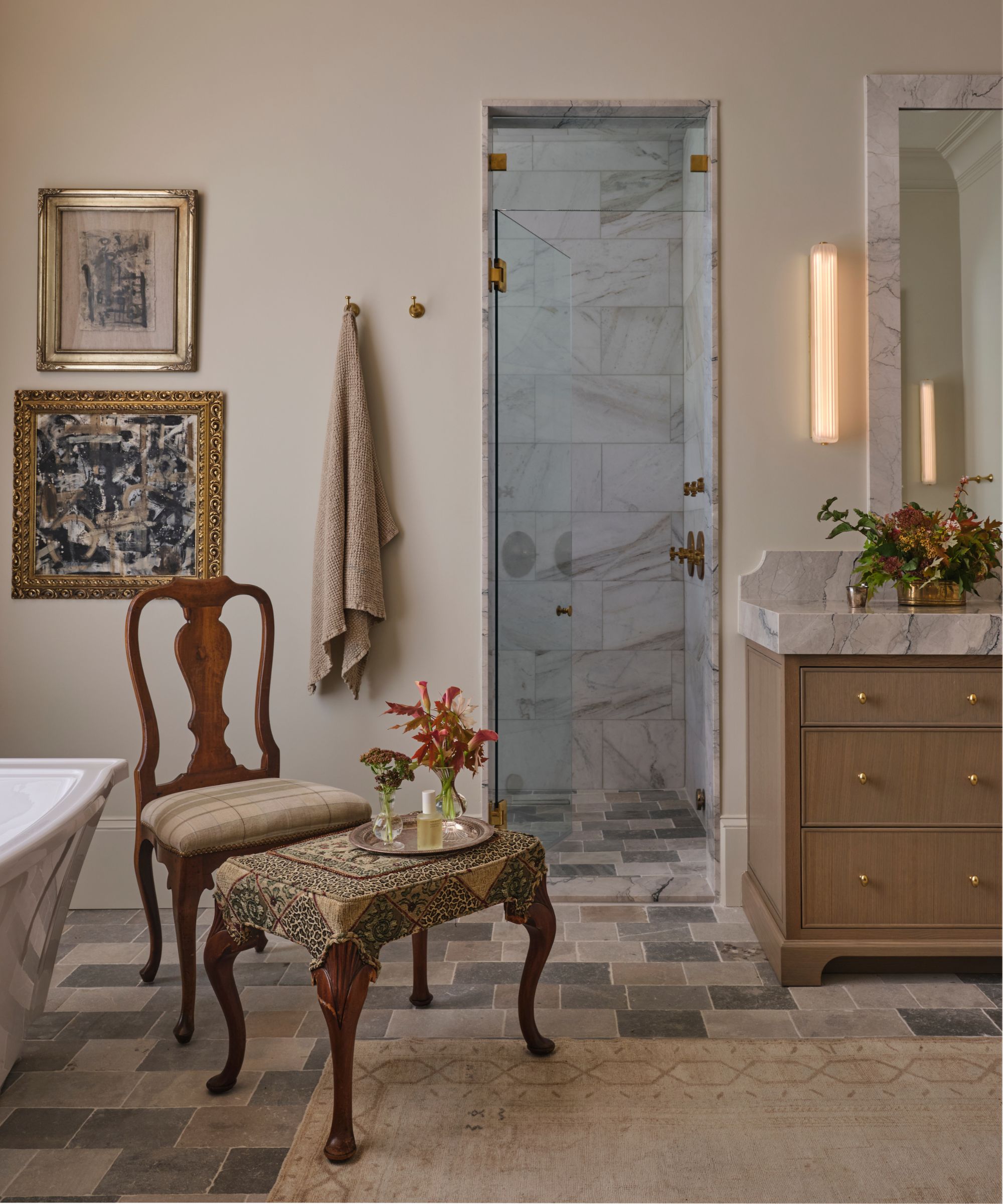 a soft neutral bathroom with stone flooring, freestanding tub, marble walk-in shower, oak vanity with solid marble countertops and antique art and furniture to style the room