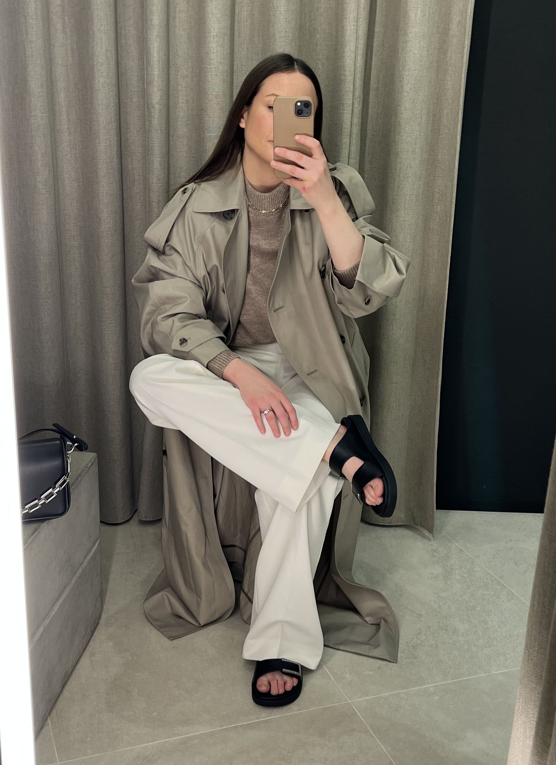 Senior Shopping Editor, Emily Dawes, wears trench coat and white trousers while posing for a selfie in the mirror.