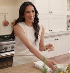 Meghan Markle wearing a white sleeveless tank and pants cooking in a kitchen with white cabinets and laughing 