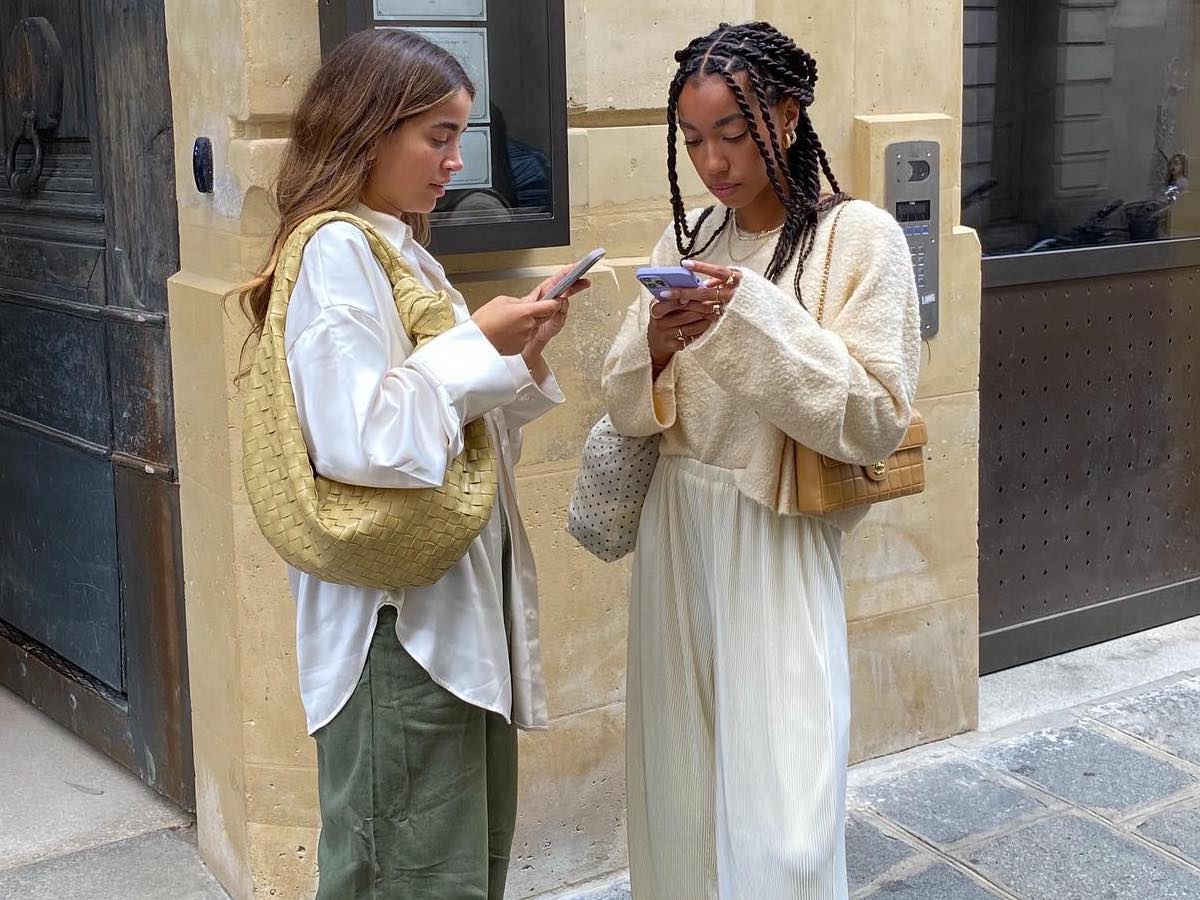 Female influencer Amaka Hamelijnck and a friend pose on the sidewalk in stylish neutral outfits while looking at their cell phones.