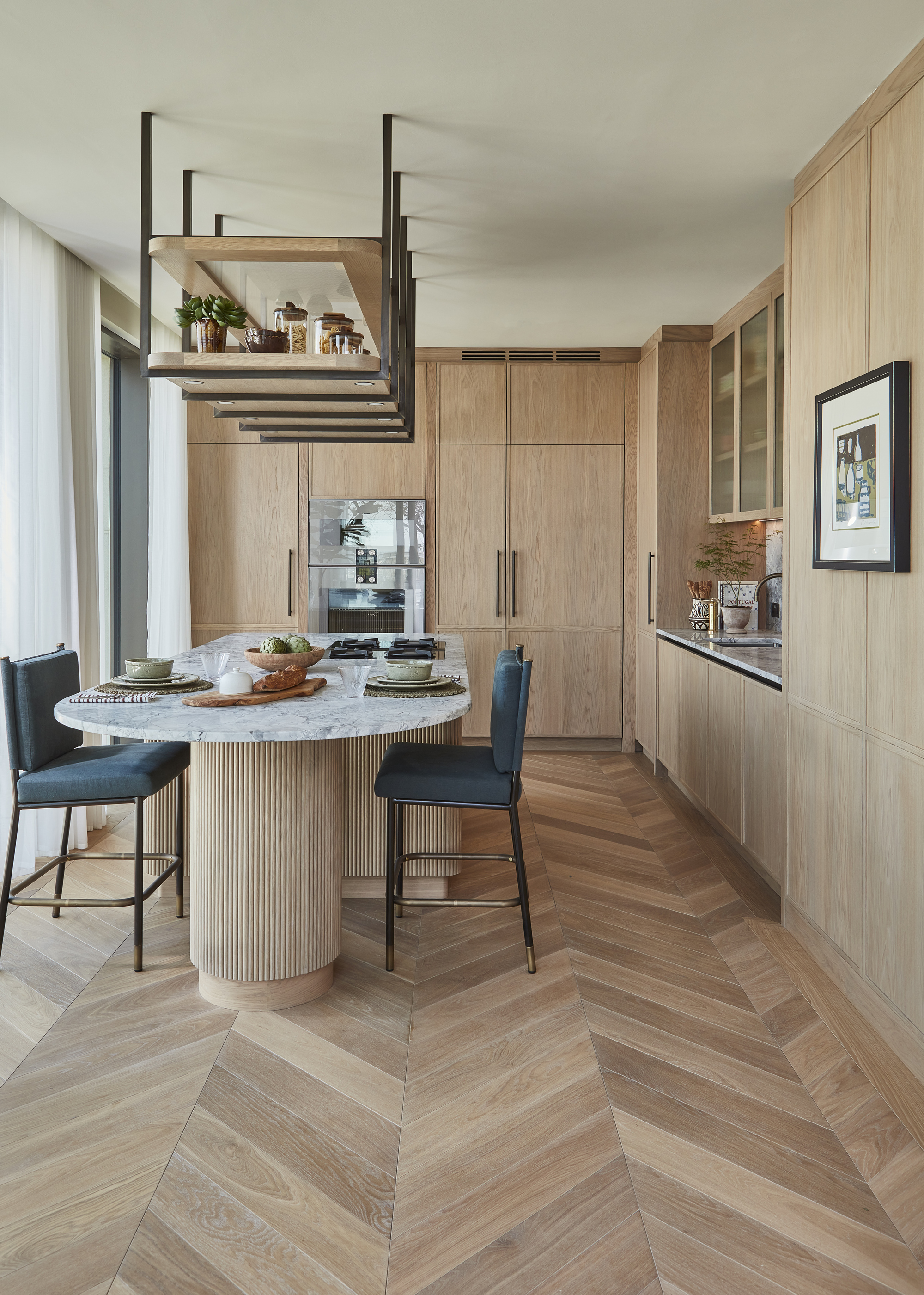 A kitchen with wooden floor-to-ceiling cabinetry, herringbone wooden flooring, and an island with a wooden fluted base and a marble counter