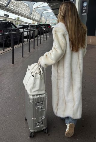 A winter travel outfit is shown in a photo of a woman standing in an airport terminal wearing a long white fur coat over a sweater and jeans styled with white sneakers, a white handbag, and silver carry-on luggage