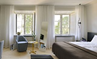 Hotel Skeppsholmen, Stockholm. A hotel room with a white double bed, a black side table, a floor lamp, a blue fabric chair, a round wooden coffee table, a striped black and white rug and two large windows with white curtains.