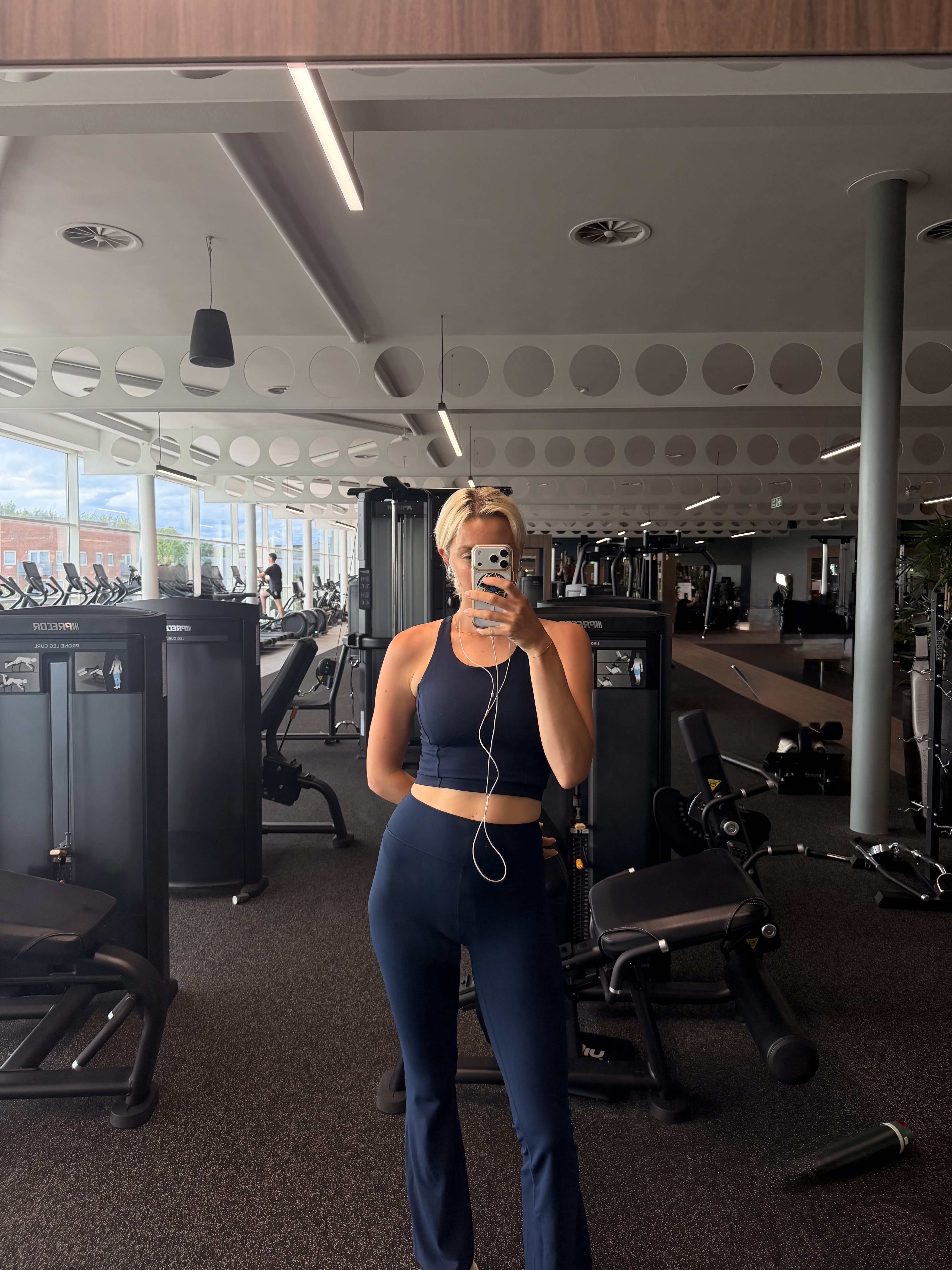 A woman with blonde hair taking a mirror selfie in a gym wearing a matching navy workout set