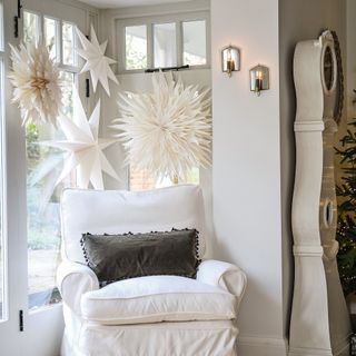 a room with a white armchair and cream grandfather clock, beside a window covered in large paper Scandi-style star decorations