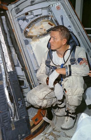 a man in a spacesuit crouches in the doorway of a small cone-shaped capsule