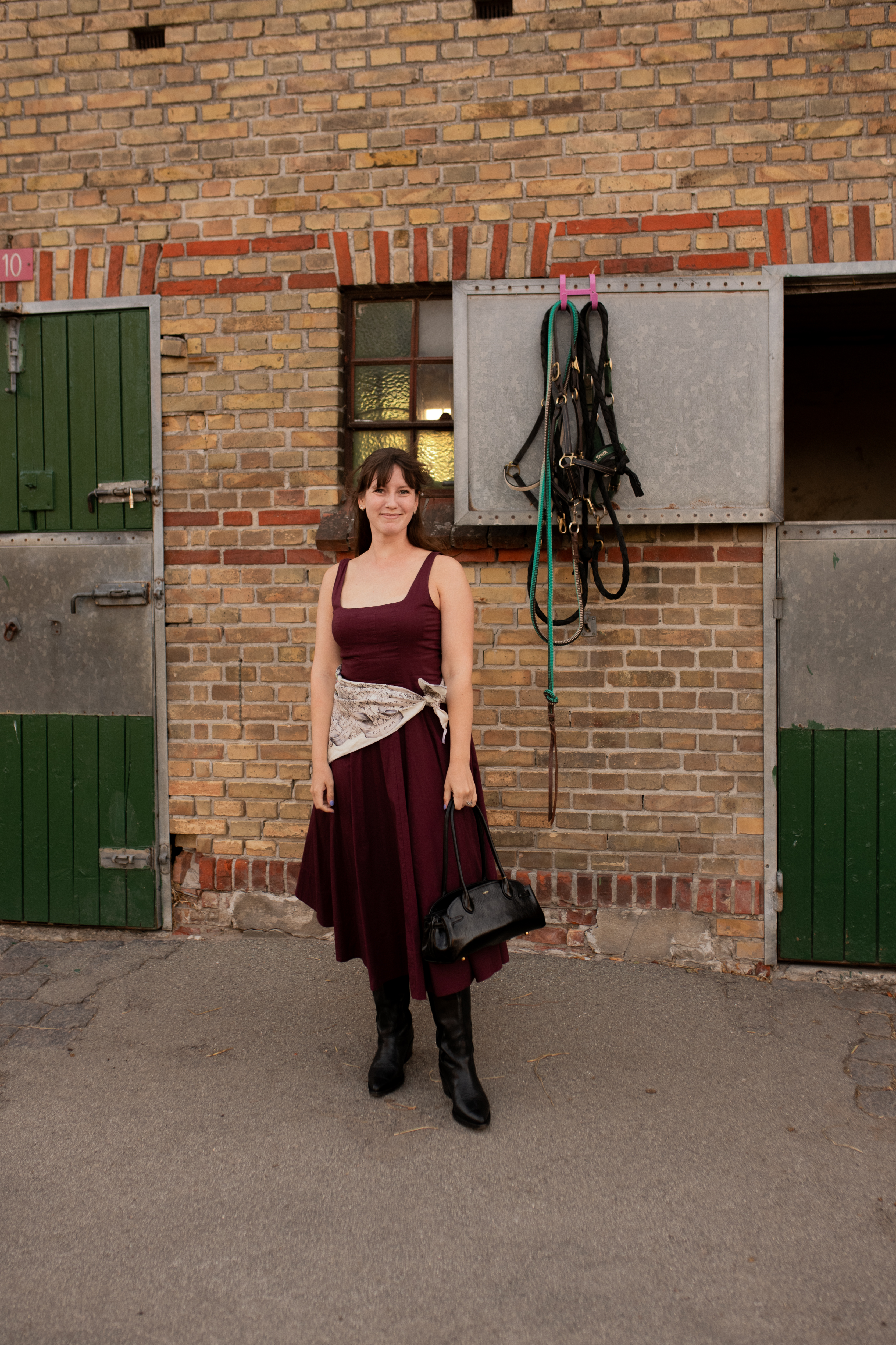 Halie LeSavage stands in front of stables wearing a tank dress with a Dior scarf and cowboy boots