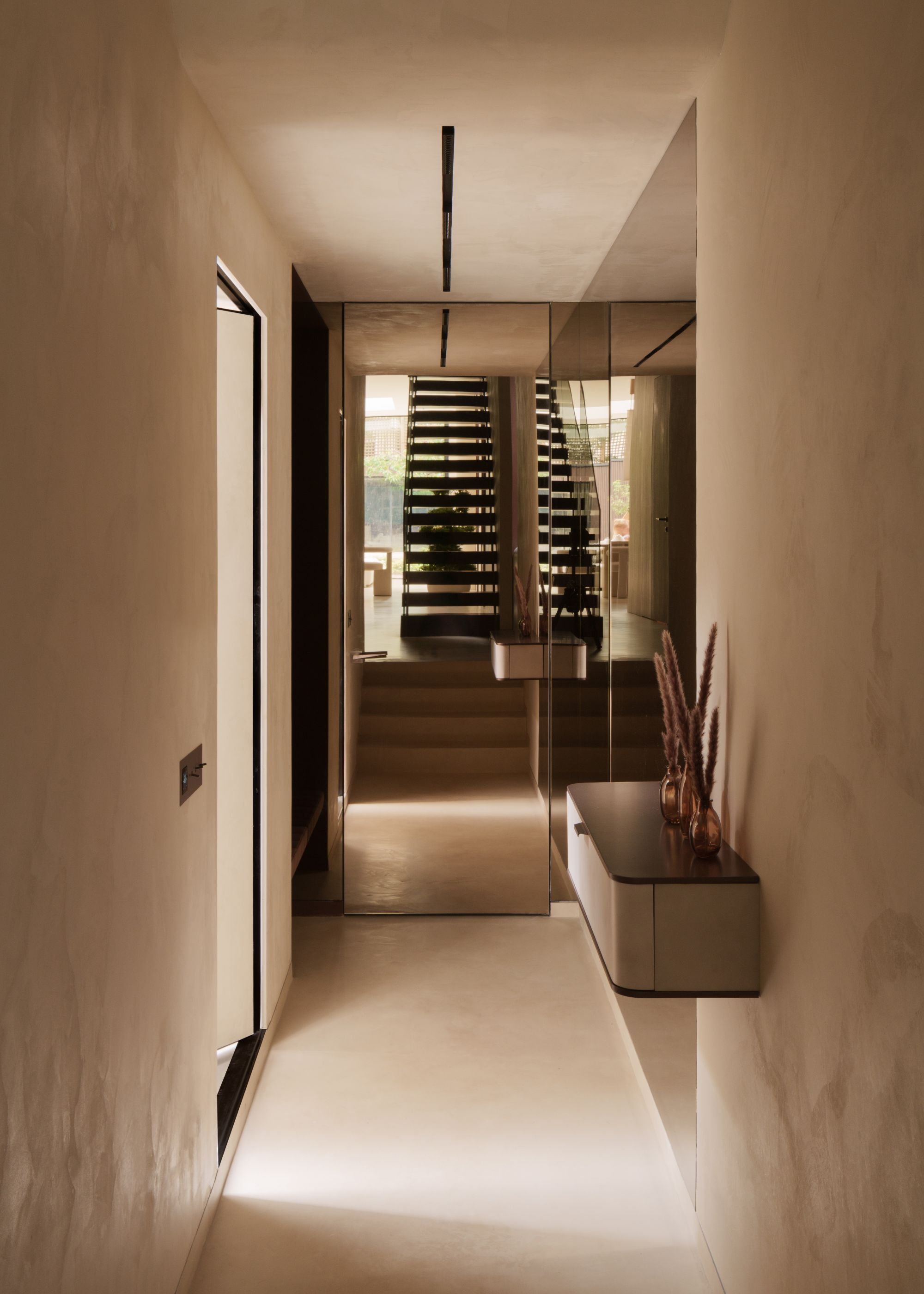 A hallway with stairs ahead with light coming through the slats, and microcement details on the walls, with a floating shelf with a display of mini vases with dried foliage