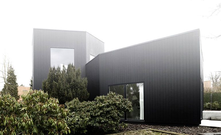 Exterior view of the villa Wienberg. The villa has two connected structures, the facade is covered with black wooden boards and is surrounded by foliage.