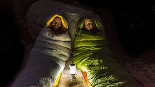 Man and a woman in sleeping bags smiling lit by a lantern at night.