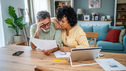 An older couple look worried as they look at paperwork at their dining room table.