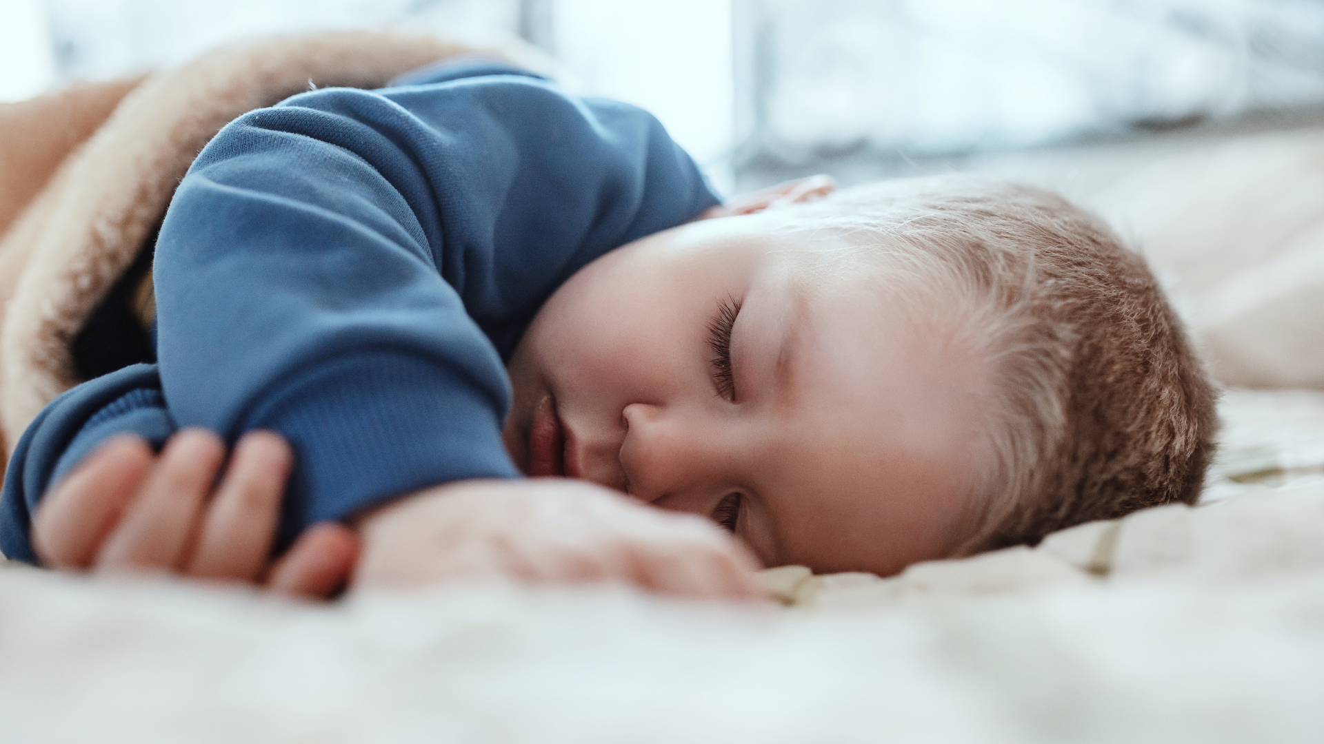 A young child has a nap during the day. He is wearing a blue sweater and is covered by a beige blanket.