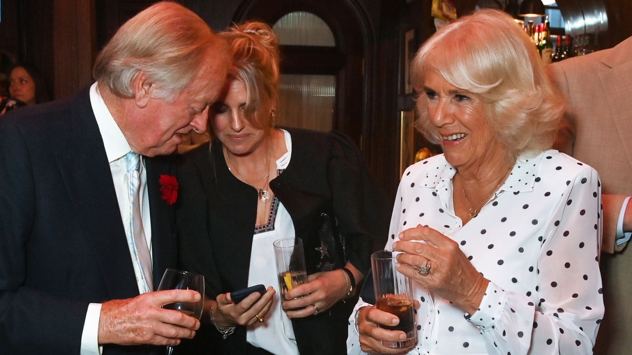 Queen Camilla wearing a white polka dot dress holding a drink and laughing standing next to ex husband Andrew Parker Bowles and daughter Laura Lopes