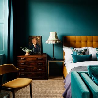how to decorate a north-facing room, teal bedroom with turmeric upholstered bed, matching retro armchair, coir carpet, vintage chest of drawers, vintage style table lamp, portrait, teal cushions, white bedding, grey/lilac blanket