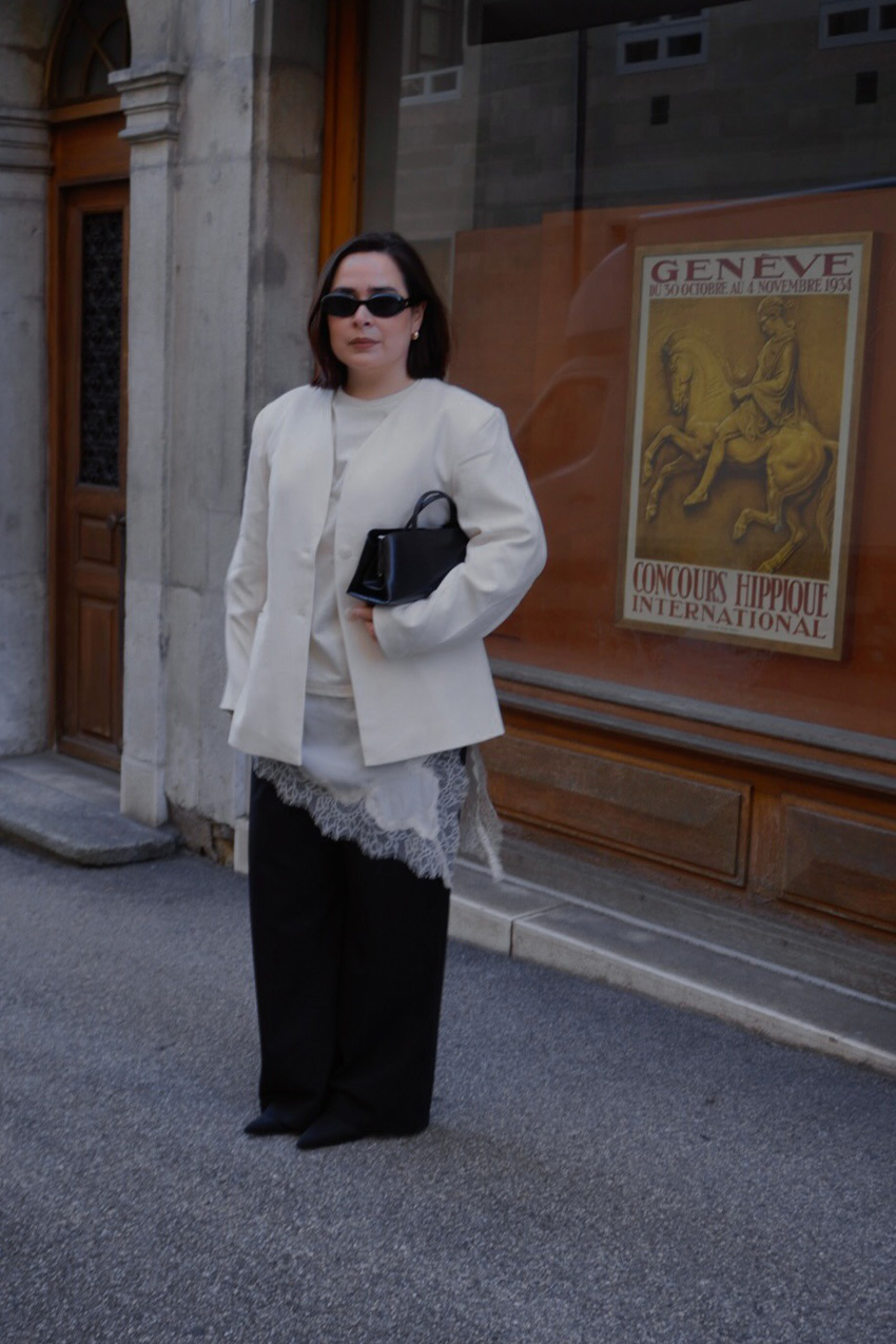 A sweater styling trend is shown in a photo of a woman&#039;s outfit, where she&#039;s standing outside in front of a storefront wearing a white blazer over a white cashmere t-shirt layered over a white slip dress with black wide-leg jeans, black pointed heels, and a black frame bag