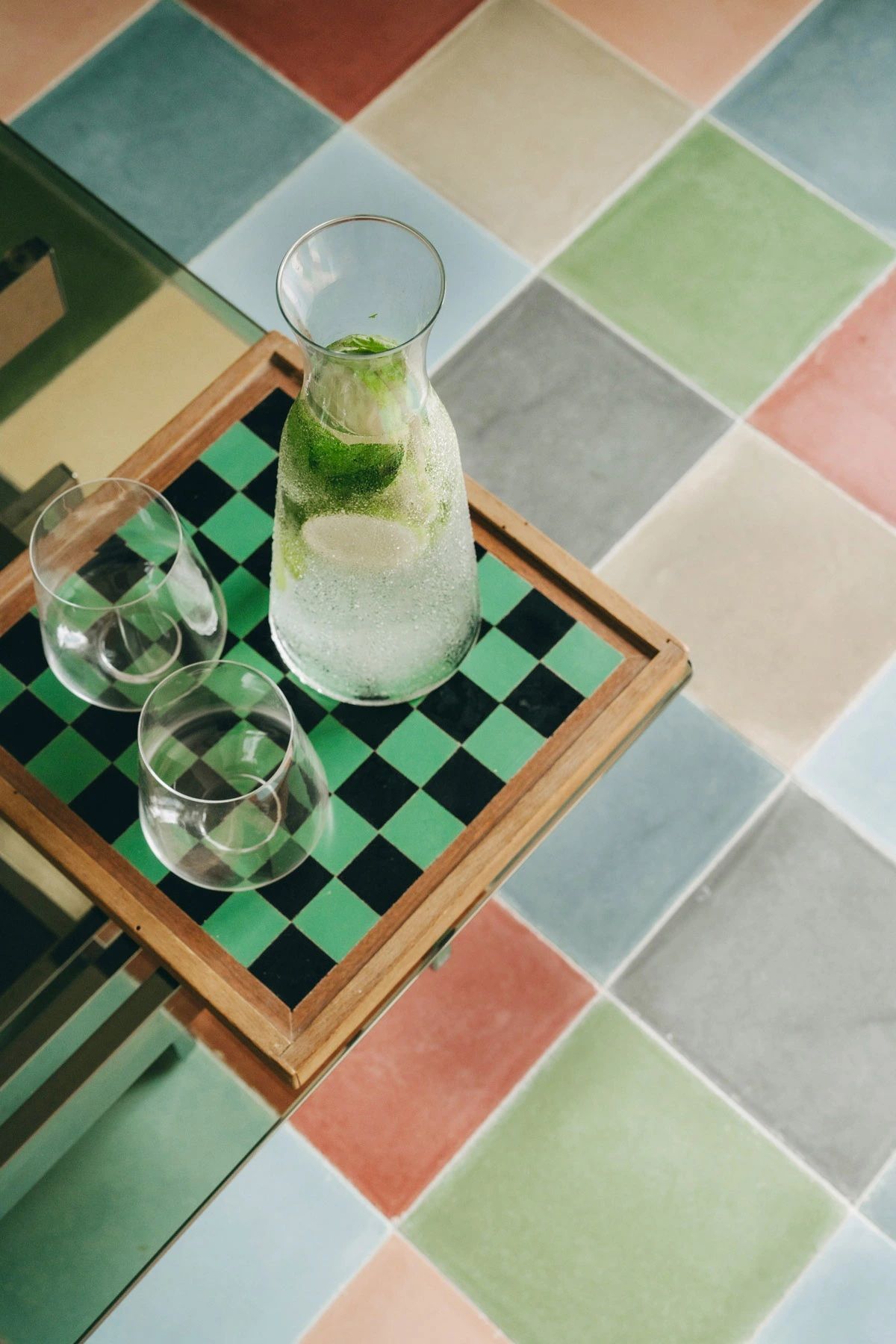 A checkered floor in pastel hues of light blue, red, green, orange, and beige serves as a backdrop to a checkered chess board onto which a pair of glasses and a water glass jug filled with lemon and mint.