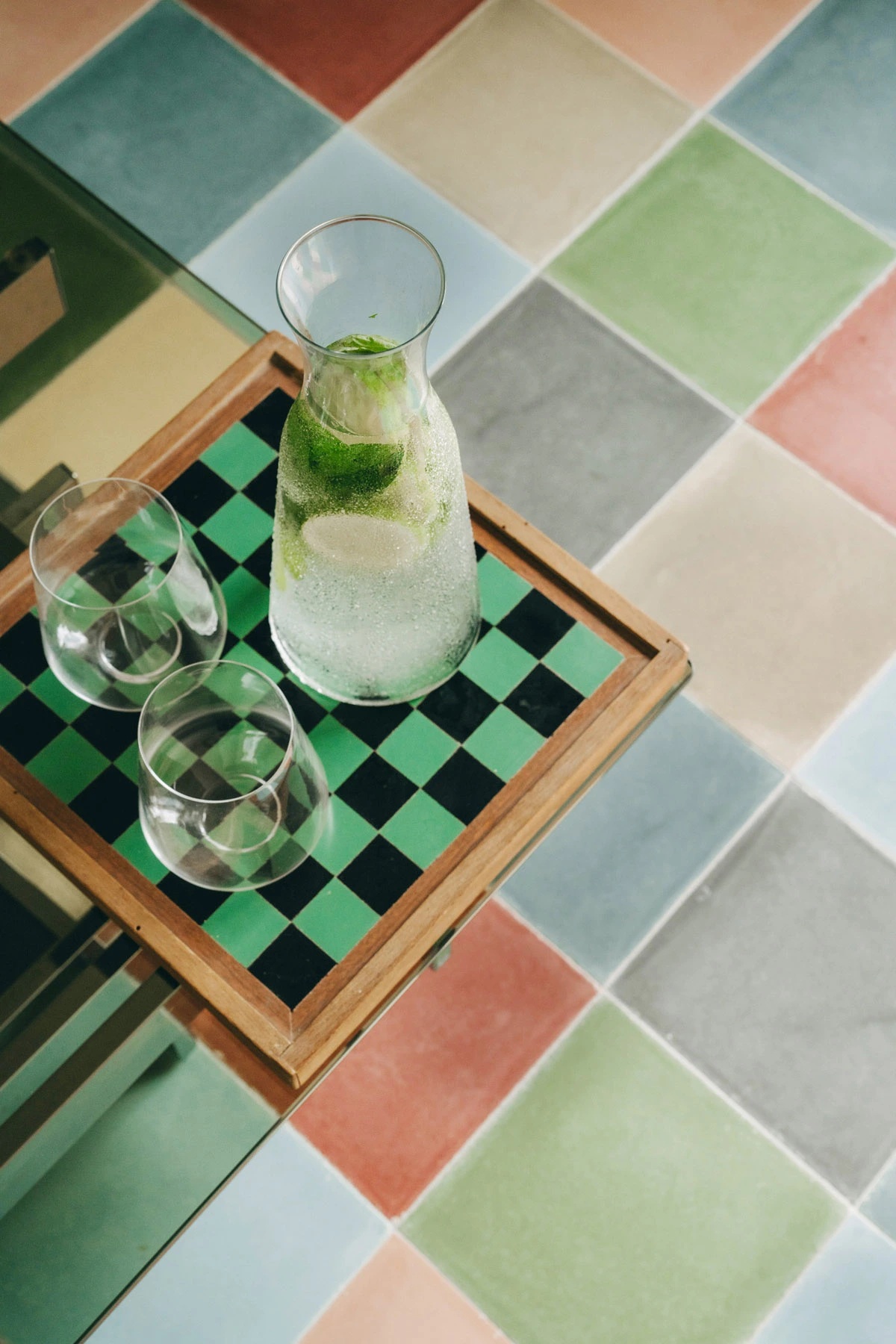 A checkered floor in pastel hues of light blue, red, green, orange, and beige serves as a backdrop to a checkered chess board onto which a pair of glasses and a water glass jug filled with lemon and mint.
