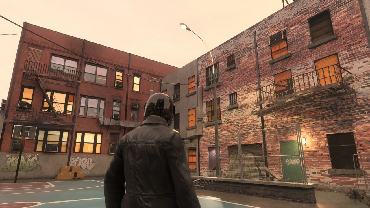 GTA IV character in a dark jacket stands facing urban buildings with a basketball court in the foreground; buildings are brick with orange windows and visible graffiti.