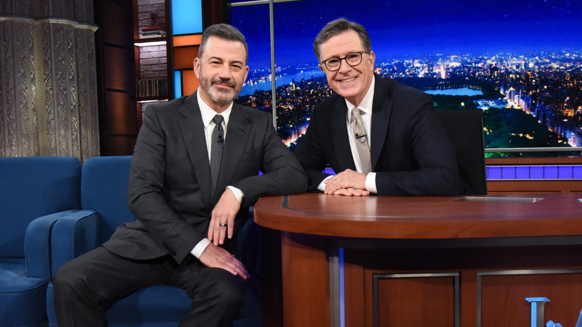 Jimmy Kimmel and Stephen Colbert sitting next to each other and smiling on The Late Show. 