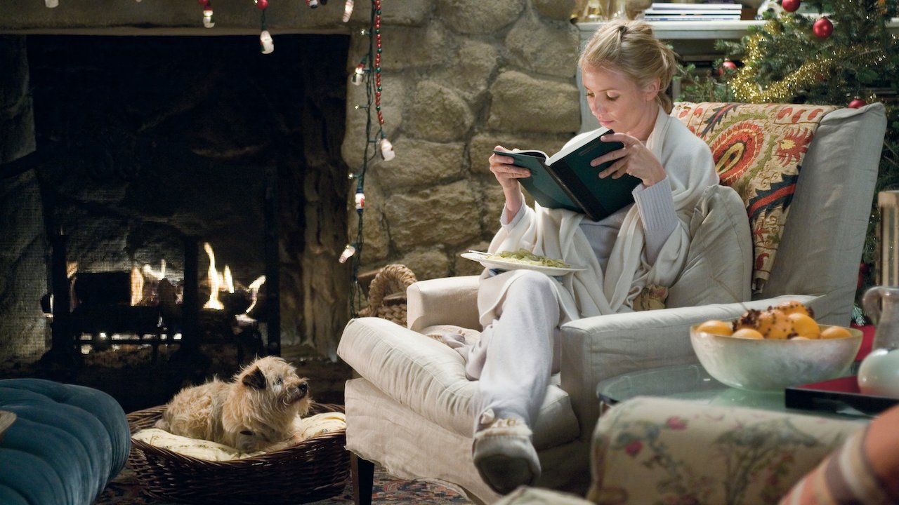 A woman in cosy loungewear sits curled up in an armchair reading a book beside a glowing fireplace, while a small scruffy terrier rests in a wicker dog bed nearby. A decorated Christmas tree glows softly behind her.