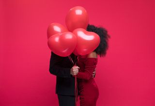 A couple celebrating Valentine's day with red heart-shaped balloons covering their faces.