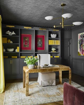 A home study with a burl wood desk and built-in shelves painted grey displaying yellow books and decor