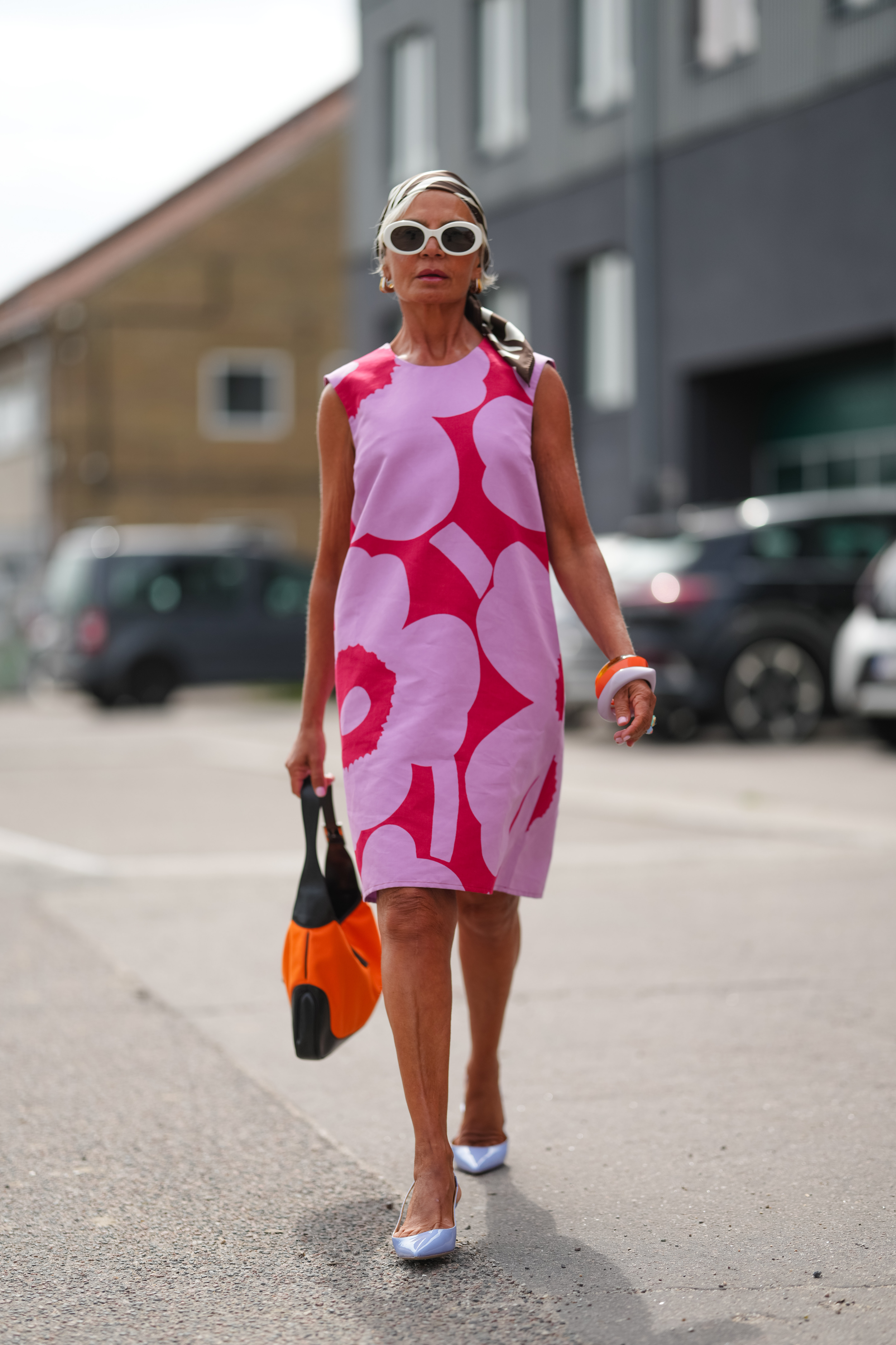 Grece Ghanem wears white oval-frame sunglasses, a black-and-white printed headscarf tied over brown-and-gray hair pulled back, and pink lipstick; a sleeveless shift dress