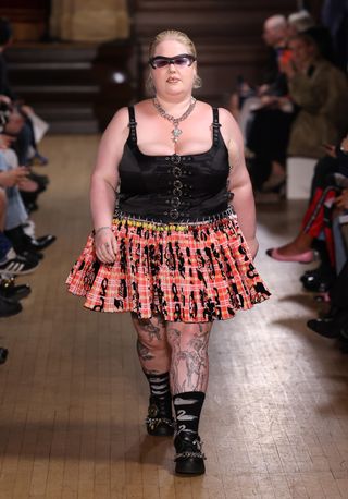 Model walking on a runway wearing a Chopova Lowena red pleated Carabiner Skirt.