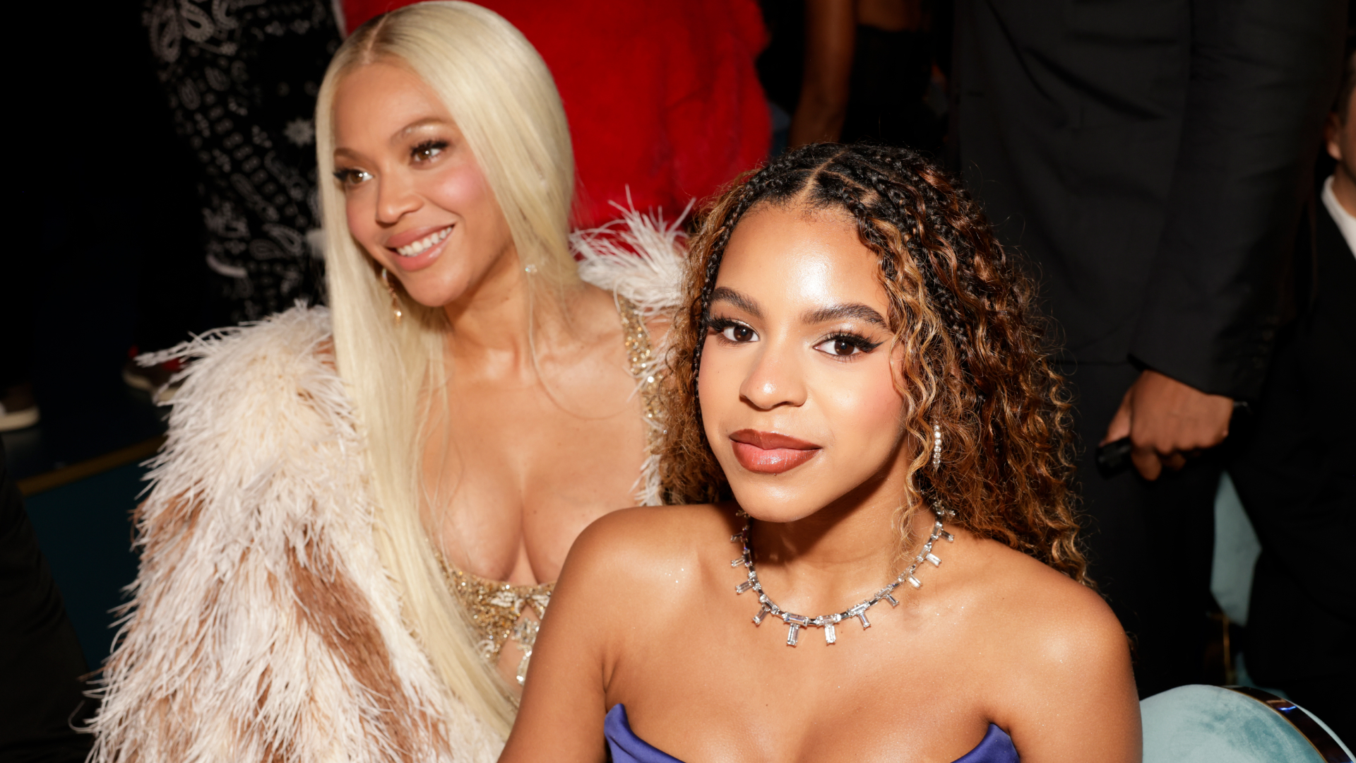 Blue Ivy wears a strapless gown and sits in front of her mom Beyonce, who has long blonde hair and is wearing a fur coat