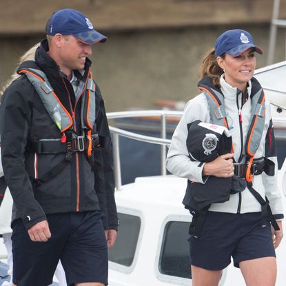Prince William and Princess Kate wearing blue baseball caps and sailing gear and shorts walking on a dock