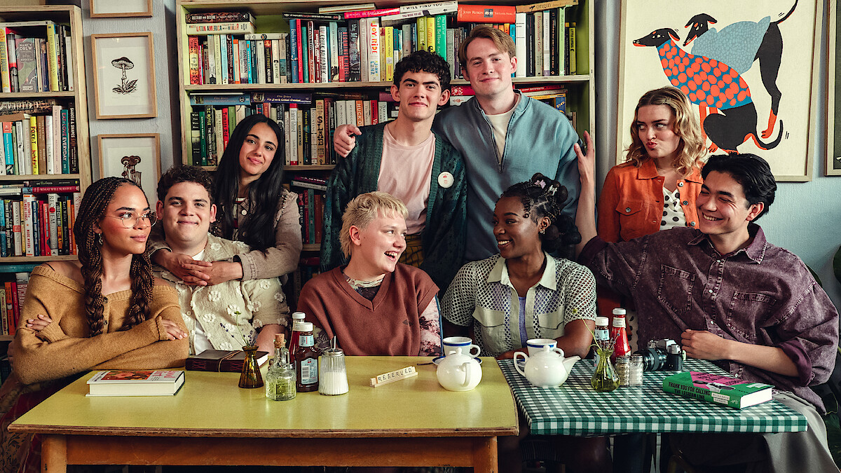 the cast of heartstopper forevever smiles posing together around two tables in a first look image