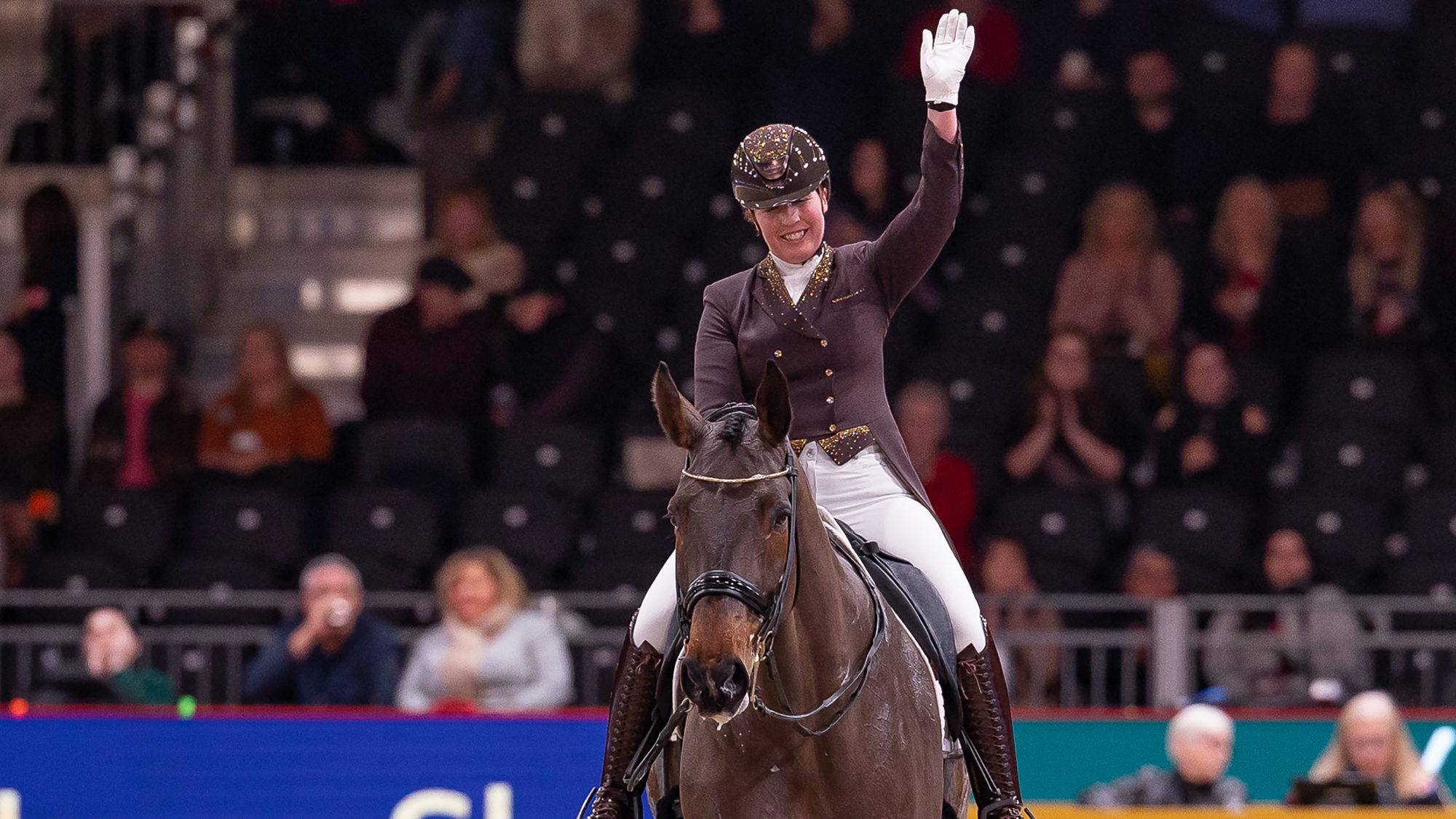 Becky Moody waves to the crowd in her signature brown jacket and tailcoat as Jagerbomb walks calmly out of the London International main area on a long rein.
