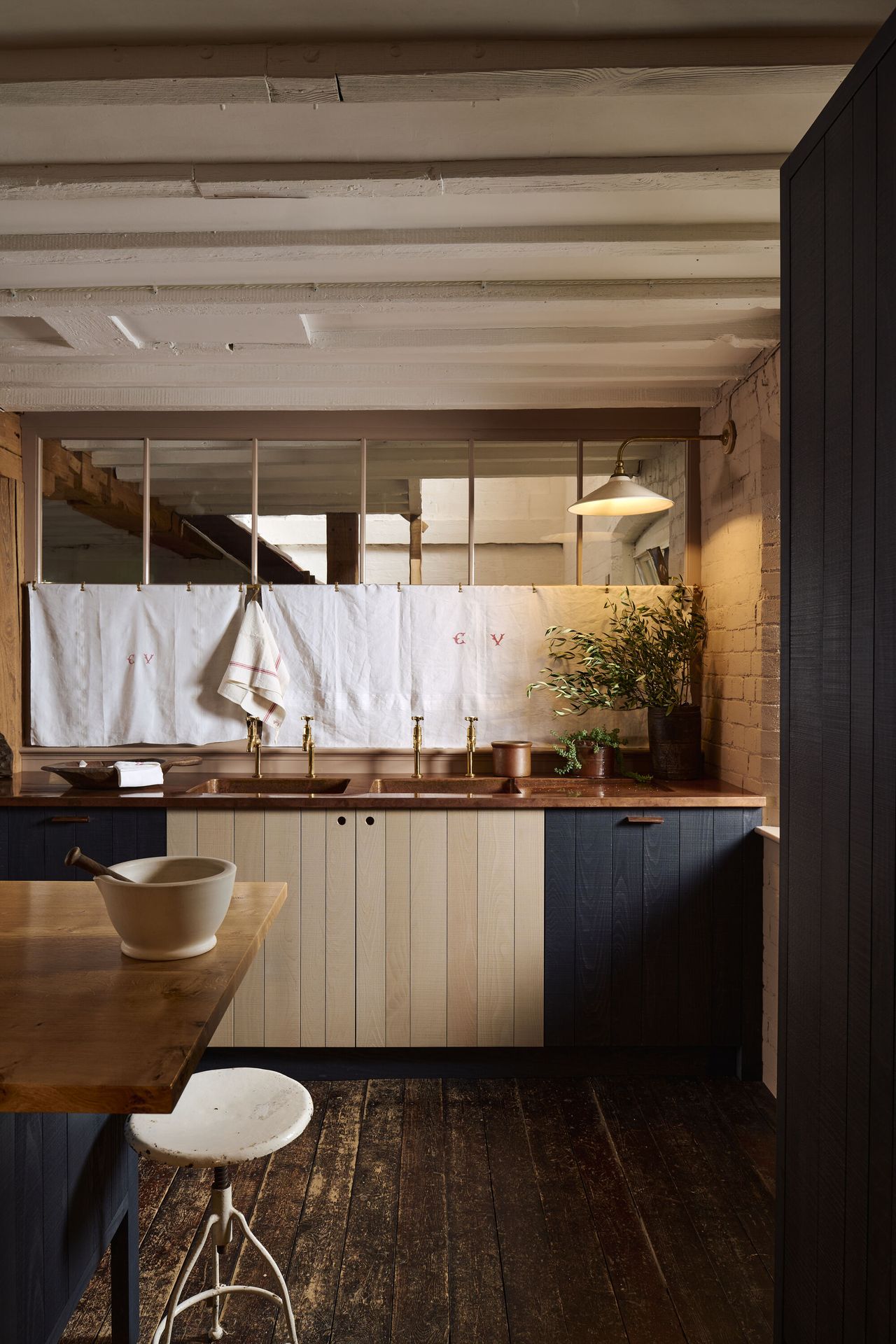 View of a kitchen countertop with an island slightly in the frame. The cabinetry is wood, stained in a white and navy color. The countertops are copper and there are indoor windows behind the sink that are covered with a white fabric.