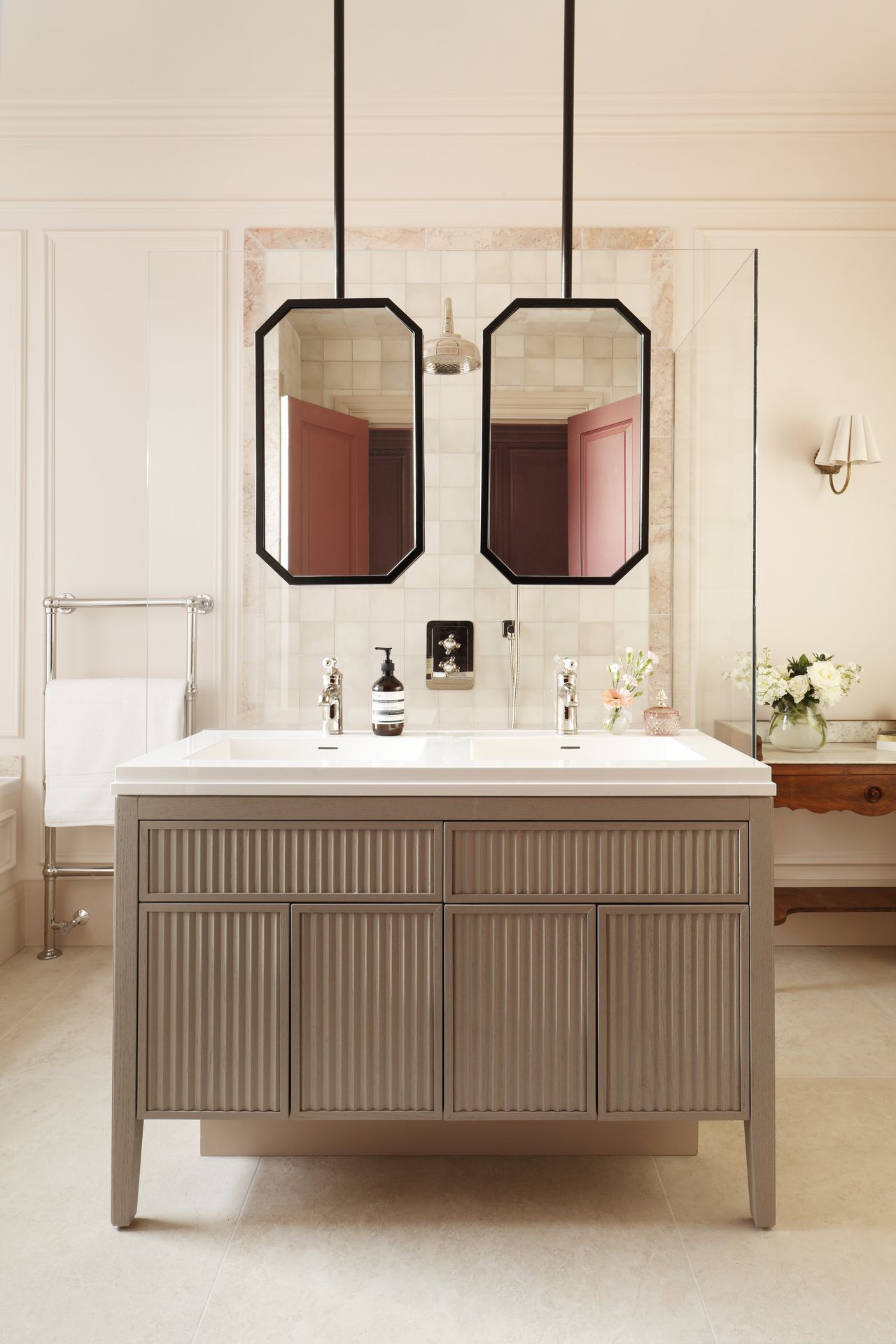 Image of a large off-white bathroom with an island in the middle that has double sinks and double suspended mirrors hanging over it.