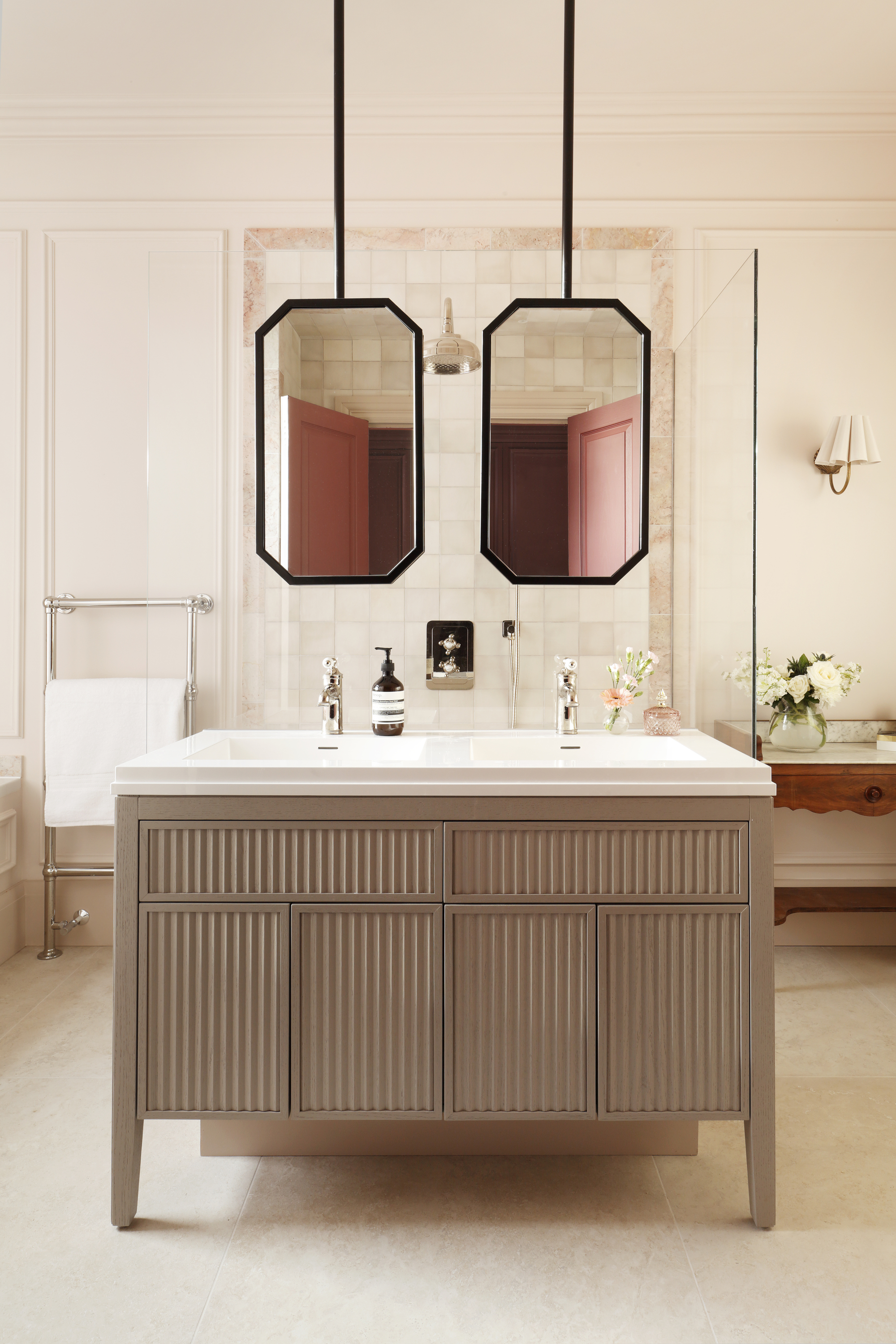 Image of a large off-white bathroom with an island in the middle that has double sinks and double suspended mirrors hanging over it.