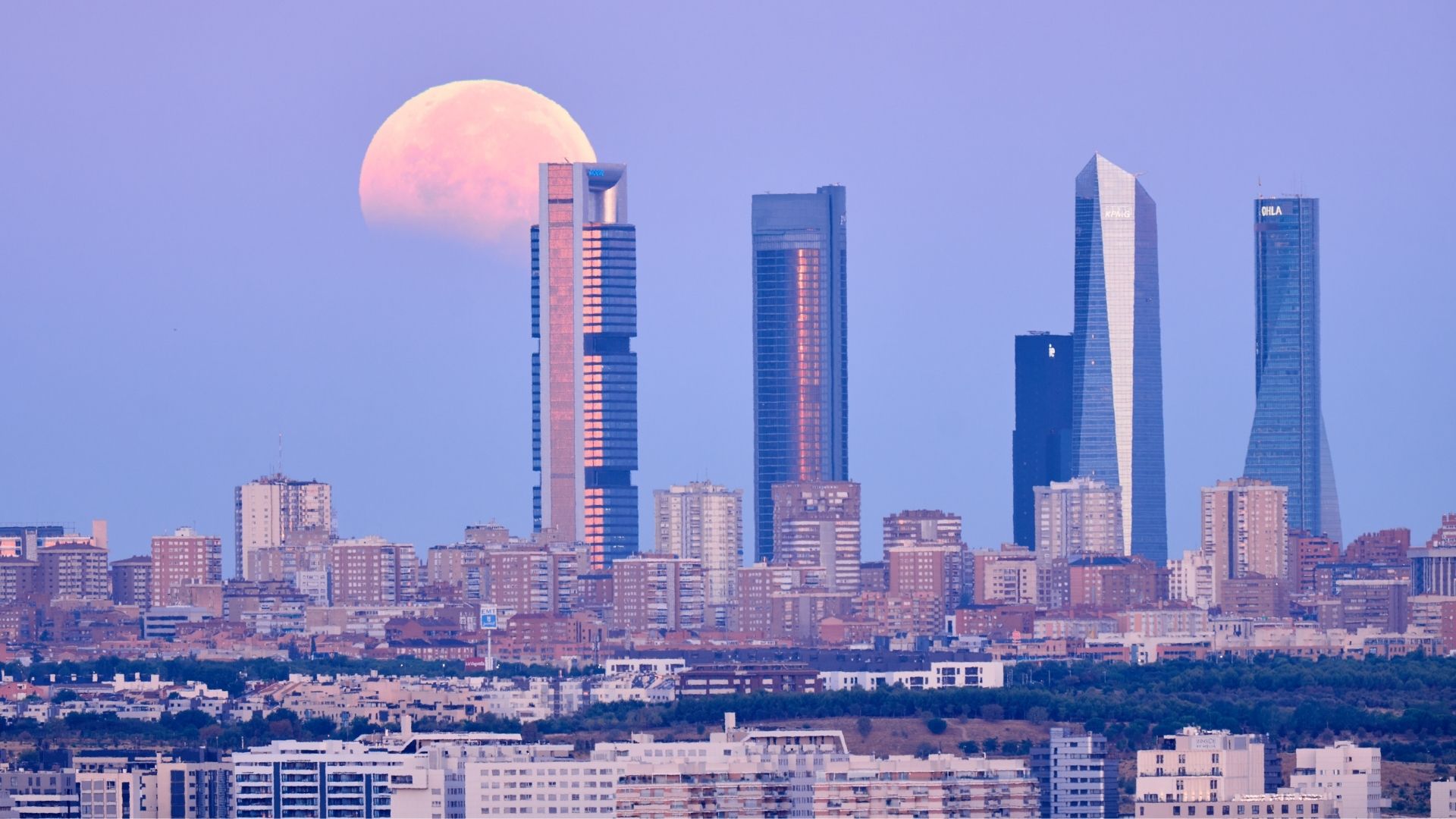 A full moon is photographed during a lunar eclipse, as Earth's shadow darkens the lunar disk above a city horizon lined with skyscrapers.