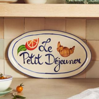 Oval serving plate with painting of an orange and a croissant and blue writing saying le petit dejeuner