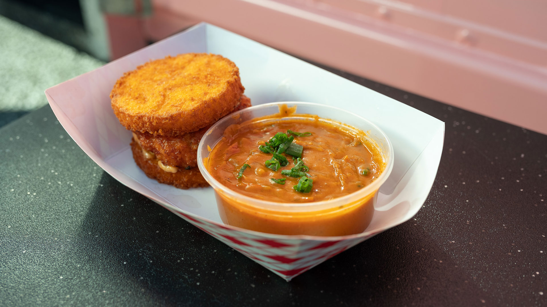 closeup of a clear plastic cup of reddish-orange gumbo, with a shrimp and grits burger on the side