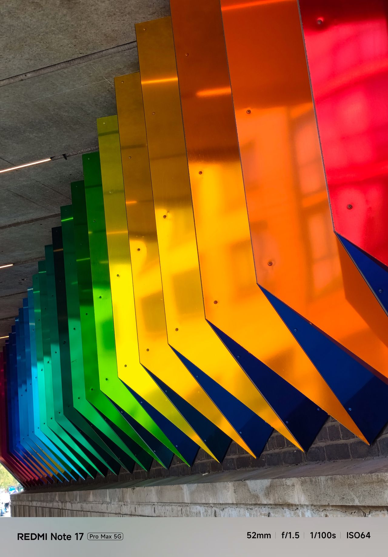 Rows of brightly colored metal panels progressing through blue, green, yellow, orange and red beneath a concrete structure.