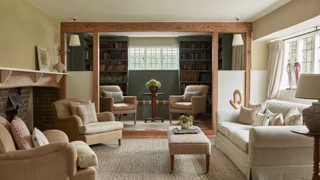 Country cottage living room with cream sofa, beige armchairs, large brick fireplace, green painted library with bookshelves and two armchairs