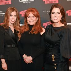 Princess Beatrice, Sarah Ferguson and Princess Eugenie posing in black dresses on the red carpet in front of a black step and repeat reading "Freedom"