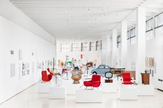A white room exhibition space with a range of furniture and objects on display including red chairs, a whicker chair, a VW car.