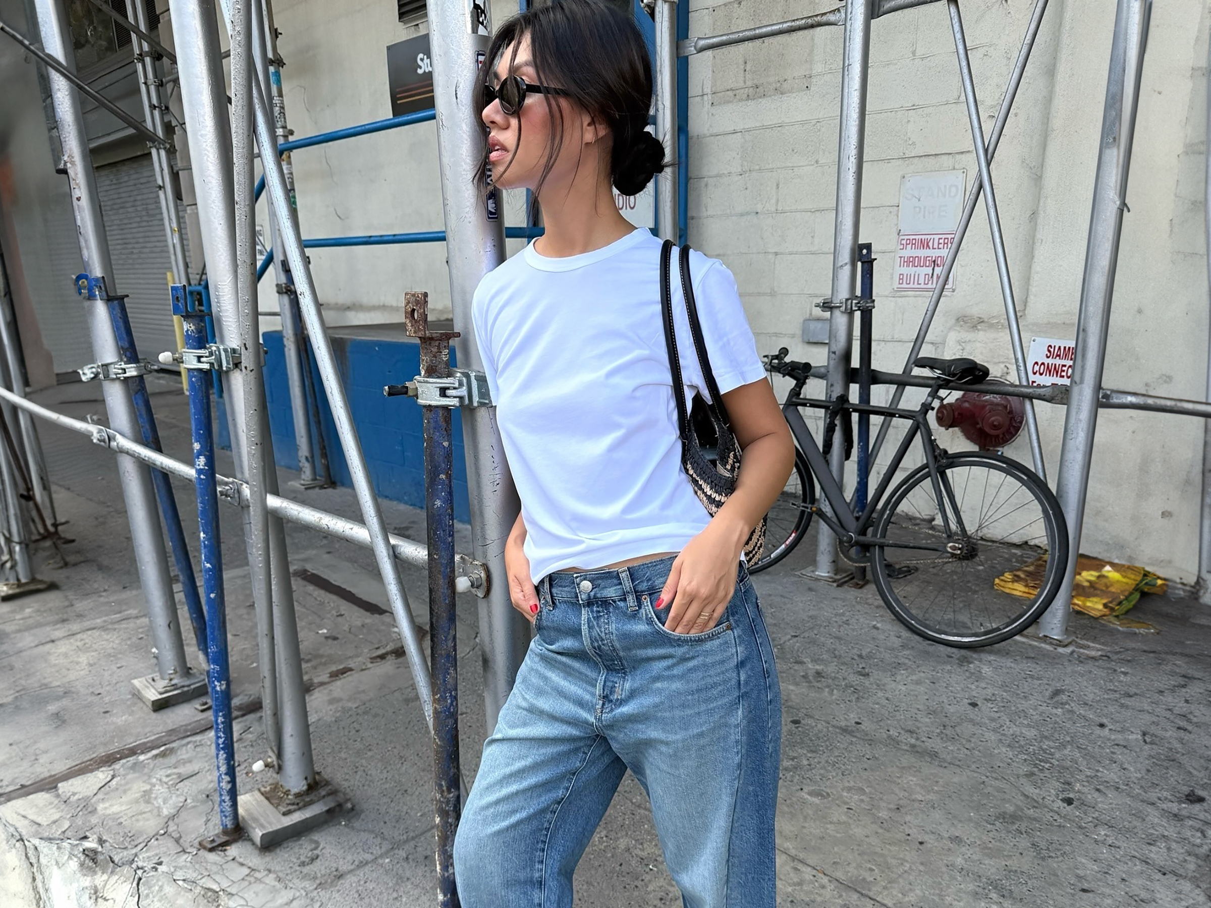Style influencer Mimi Nguyen poses on a NYC sidewalk wearing black sunglasses, a white T-shirt, black shoulder bag, and baggy Madewell jeans.