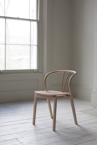Ercol ’Flow Chair’, bespoke design neutral colour wooden chair, white wash wooden floor panels, white wall room with high skirting boards, window to the left with gold fixings letting daylight in to cast shadows