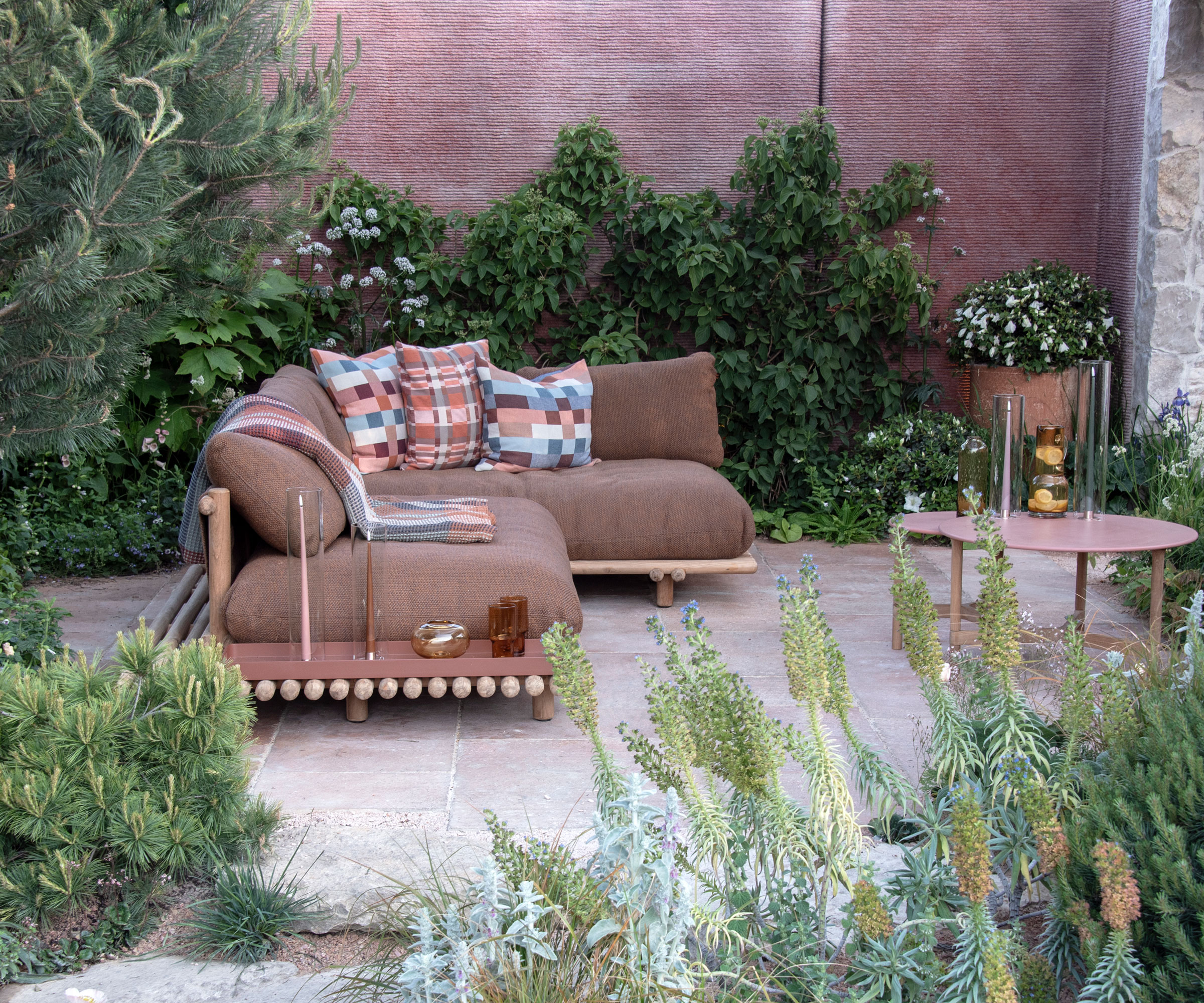 slouchy brown outdoor sofa set with geometric patterned cushions, and small garden table, on patio area surrounded by wall with climbing plants and flower beds with naturalistic planting