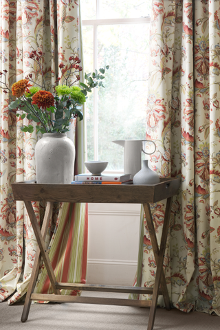 Blendworth curtain ideas. Close up of floral patterned curtains, wooden side table with white ornaments and jug of flowers