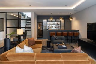basement living room/game room with large russet velvet sectional, tv, bar with bar stools, black console table, leather footstool, crittall doors to upstairs
