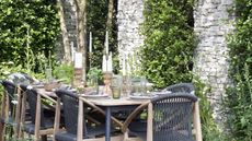 A table with nine dining chairs around it, set with candles, glassware and crockery, in a walled garden with lush greenery